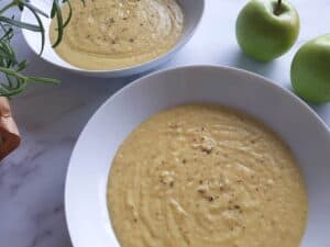 Apple and parsnip soup in two bowls with green apples in the background and fresh herbs to the side, on a marble table.