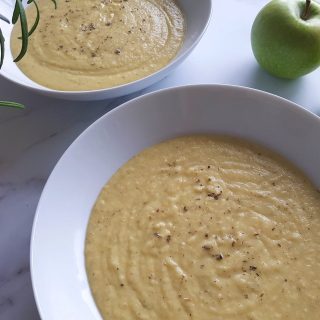 Apple and parsnip soup in two bowls with green apples in the background and fresh herbs to the side, on a marble table.