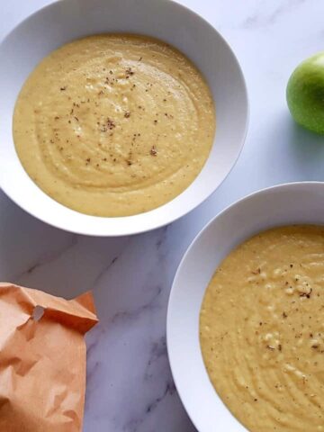 Apple and parnsip soup in white bowls with two green apples in the background and fresh herbs in the foreground.