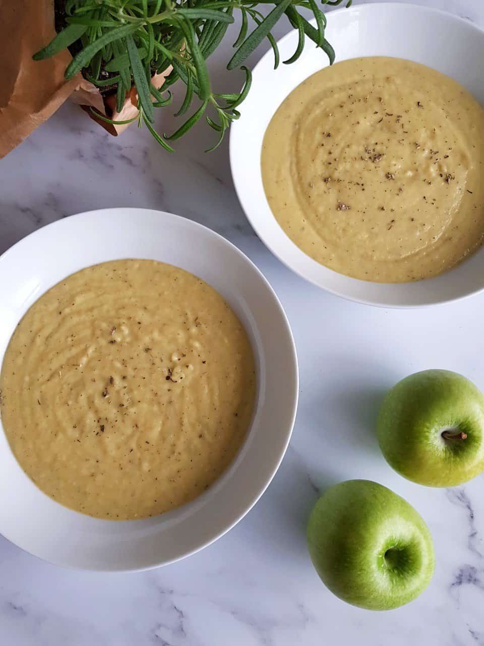 Bowls of apple and parsnip soup, with green apples in the foreground and fresh herbs in the background.