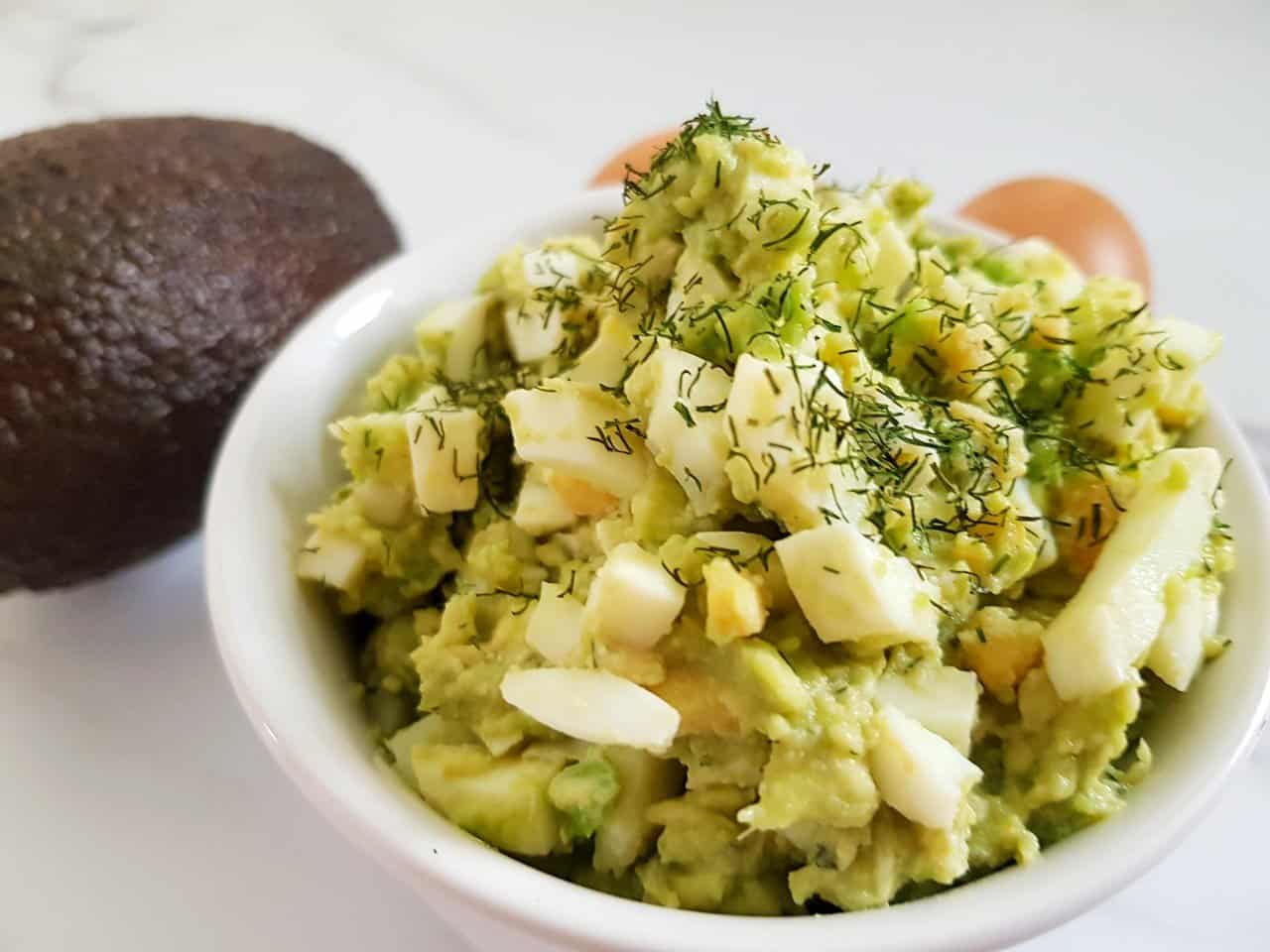 Avocado egg salad in a white bowl with eggs and avocado in the background.