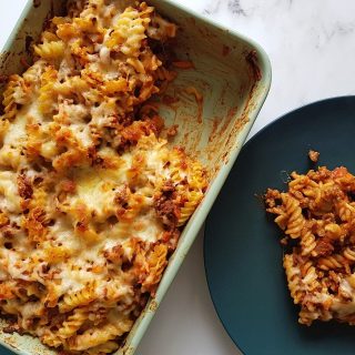 Beef pasta bake in a casserole dish with a plated portion on the side, on a marble table.