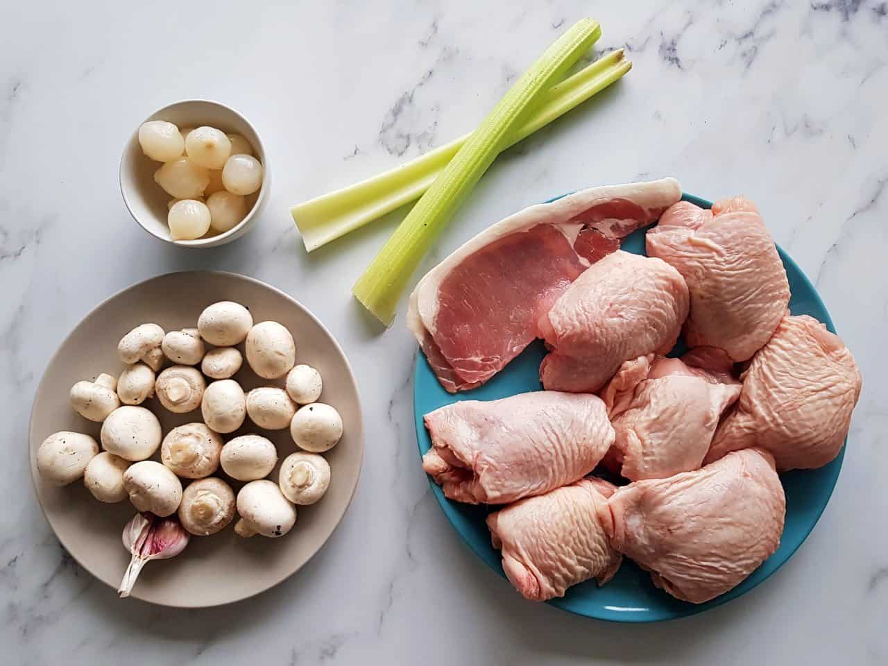 Raw ingredients for coq au vin on plates: silverskin onions, mushrooms, celery, bacon and chicken thighs.