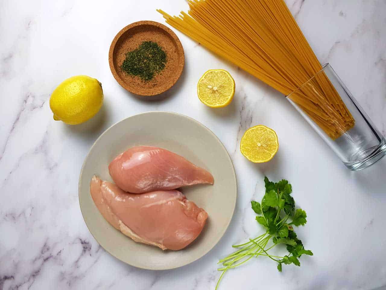 Chicken fillets, dill, lemon, coriander and pasta laid down on a marble table.