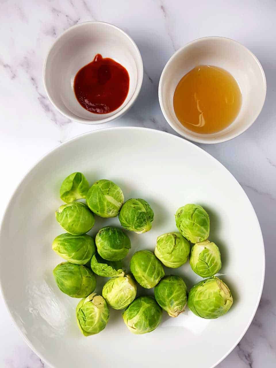Brussels sprouts on a white plate with bowls of honey and sriracha in the background.