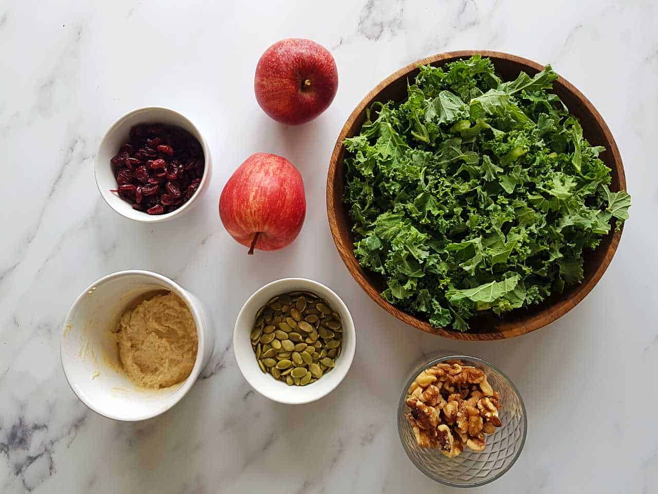 All ingredients for kale salad with apples laid out in small bowls on a marble table.