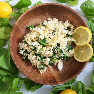 Orzo salad in a wooden bowl, surrounded by lemons and spinach leaves.