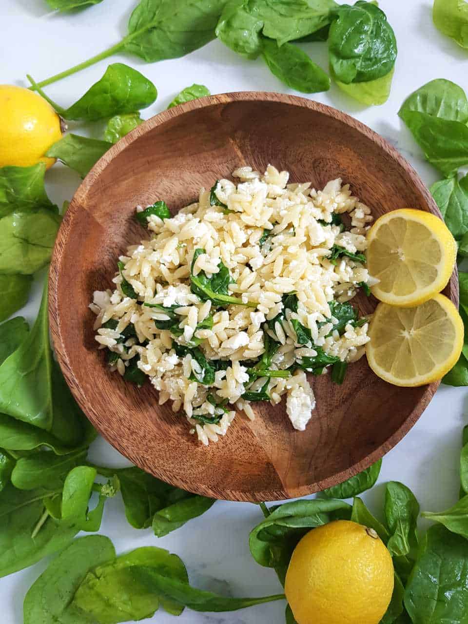 Orzo salad in a wooden bowl, surrounded by lemons and spinach leaves.