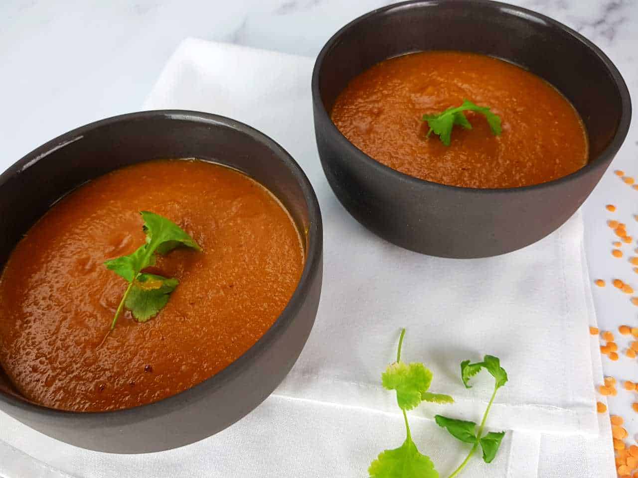 Spicy red lentil soup in black bowls on a marble table with coriander, a white napkin and lentils in front.