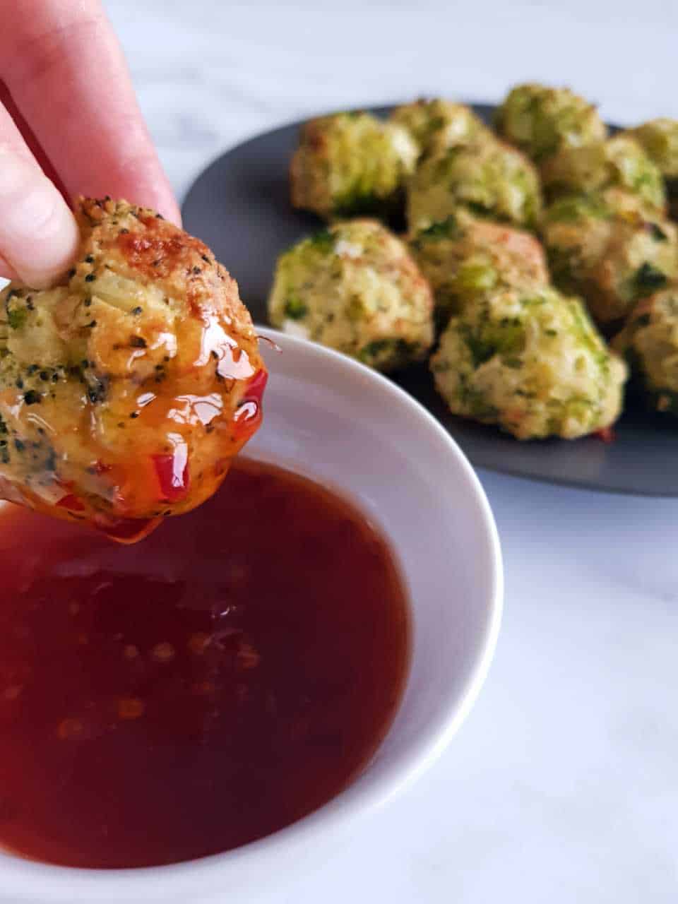 Broccoli and cheese ball dipped in a bowl of red sauce. Plate of broccoli and cheese balls in the background.