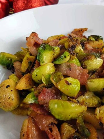 Brussels sprouts with bacon on a white plate with red baubles in the background.