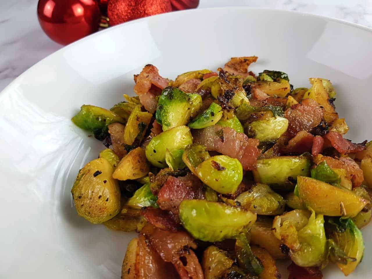 Brussels sprouts with bacon on a white plate with red baubles in the background.