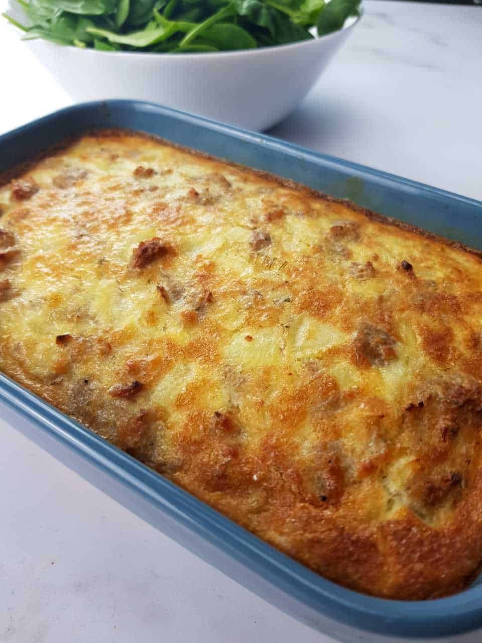 Egg and sausage casserole in a blue baking dish on a marble table. White bowl with salad in the background.