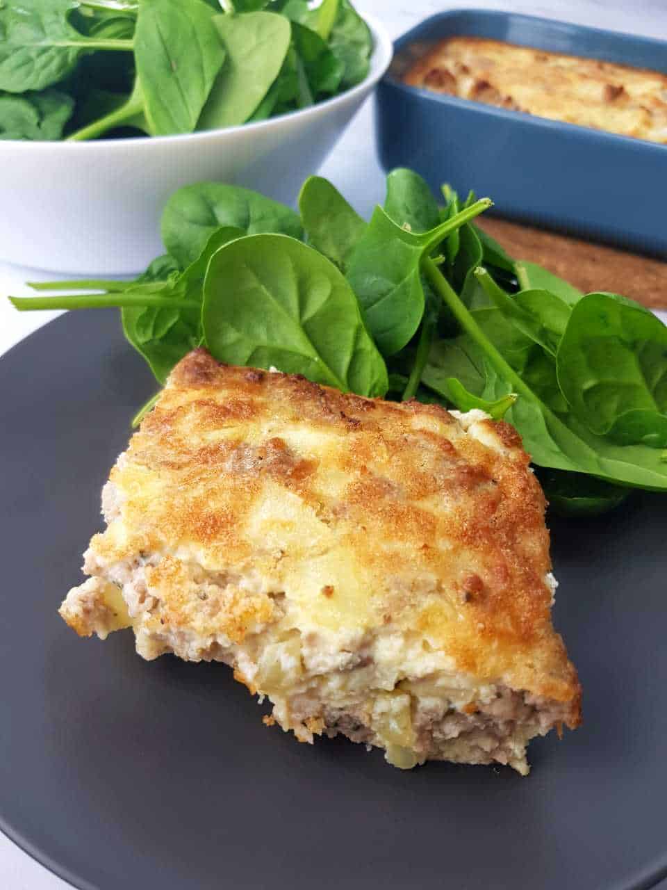 Egg sausage casserole on a gray plate with spinach. Salad bowl and blue casserole dish in the background.