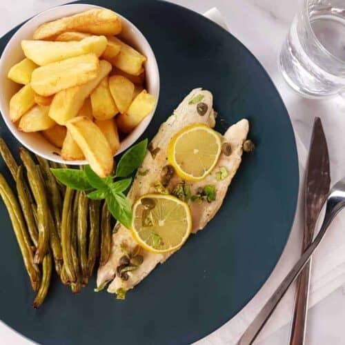 Pan seared sea bass with lemon and capers on a blue plate with chips and green beans on a marble table. Glass of water and cutlery on the side.