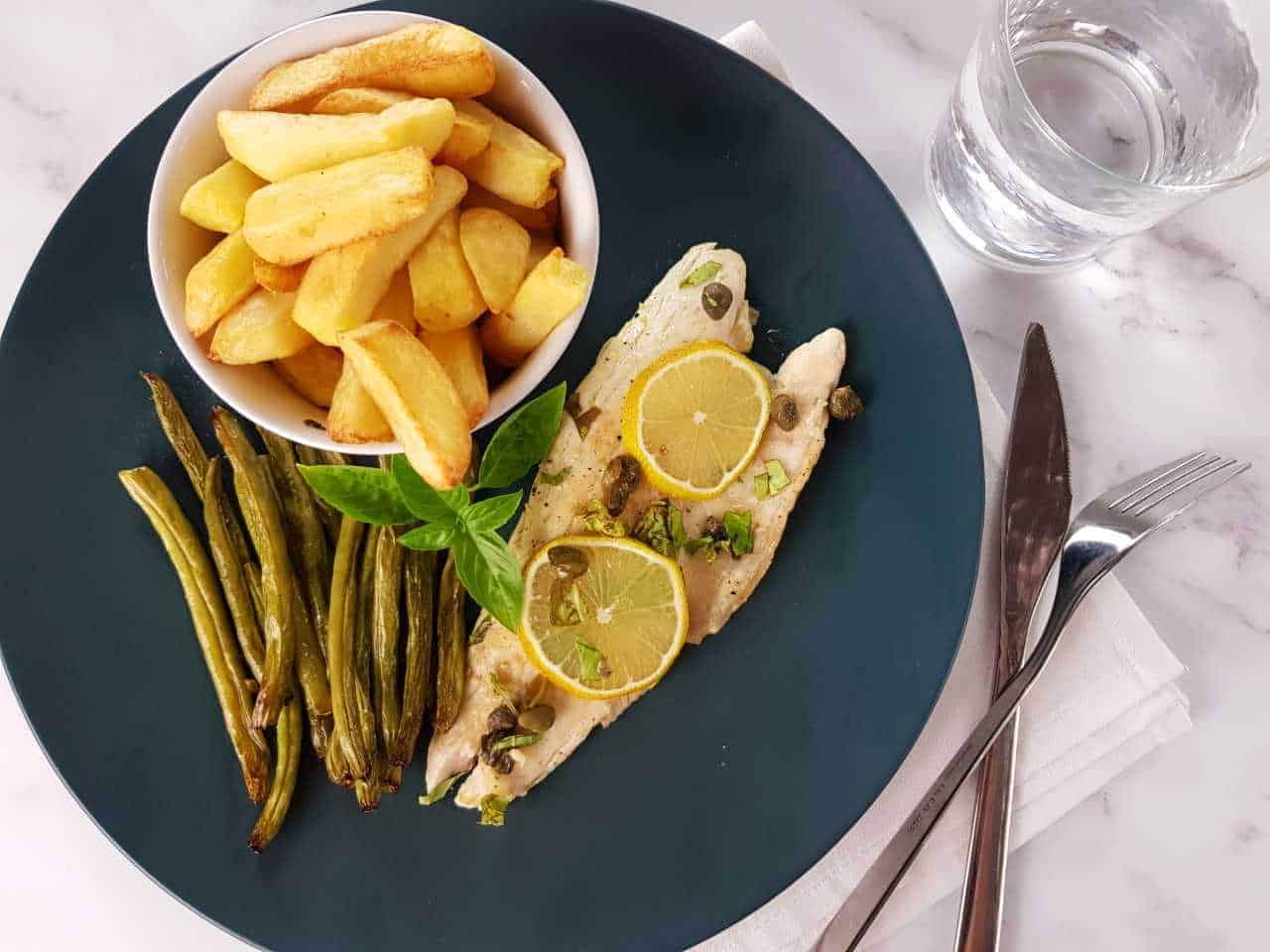 Pan seared sea bass with lemon and capers on a blue plate with chips and green beans on a marble table. Glass of water and cutlery on the side.