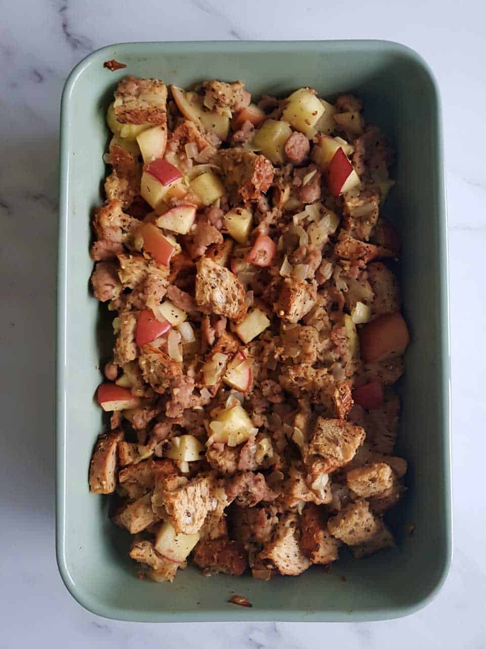 Sausage and apple stuffing in a green casserole dish on a marble table.