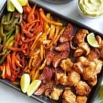 Sheet pan fajitas on a marble table with guacamole, cilantro and lime wedges on the side.