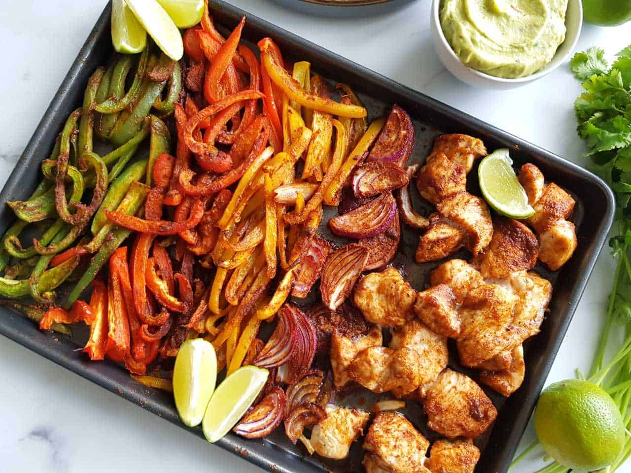 Sheet pan fajitas on a marble table with guacamole, cilantro and lime wedges on the side.