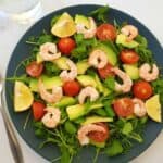 Shrimp and avocado salad on a blue plate on a marble table with a fork and a glass of water next to it.