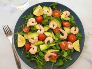Shrimp and avocado salad on a blue plate on a marble table with a fork and a glass of water next to it.