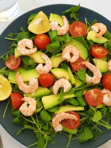 Shrimp and avocado salad on a blue plate on a marble table with a fork and a glass of water next to it.