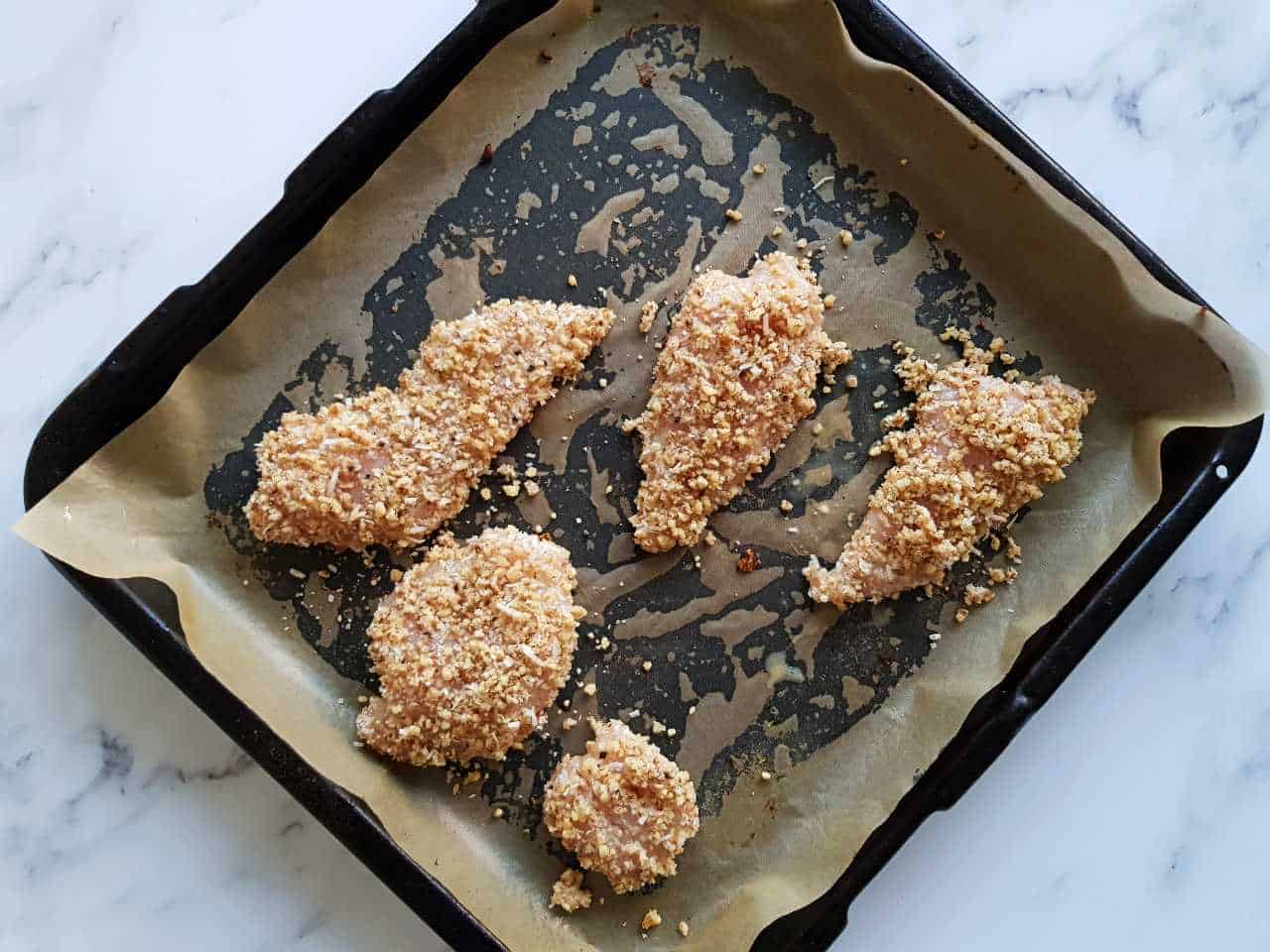Uncooked walnut crusted chicken on a baking tray on a marble table.