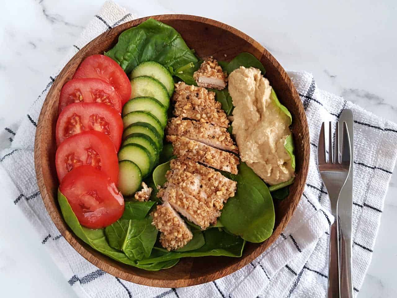 Walnut crusted chicken in a salad in a wooden bowl with cutlery and a napkin next to it.