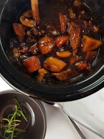 Beef and ale stew in the slow cooker on a marble table, with gray bowls and forks in front.