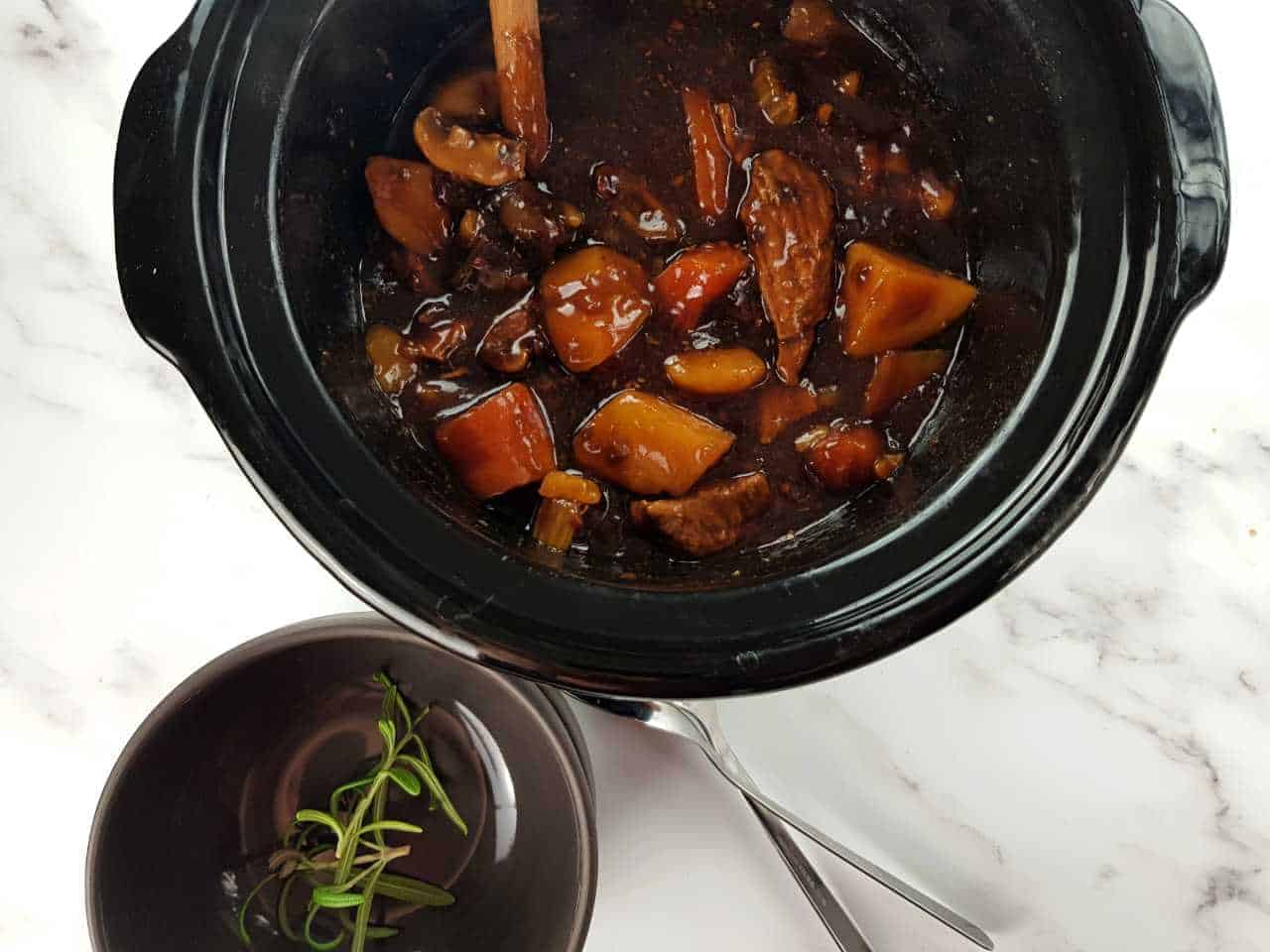 Beef and ale stew in the slow cooker on a marble table, with gray bowls and forks in front.