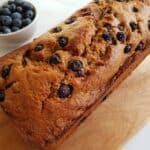 Banana & blueberry bread on a wooden board on a marble table with a bowl of blueberries on the side.