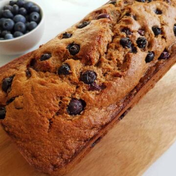 Banana & blueberry bread on a wooden board on a marble table with a bowl of blueberries on the side.