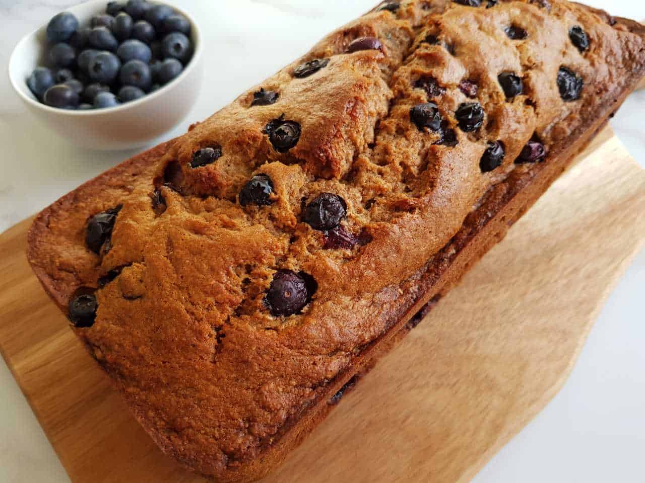 Banana & blueberry bread on a wooden board on a marble table with a bowl of blueberries on the side.