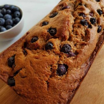 Banana bread with blueberries on a wooden board with a bowl of blueberries on the side.