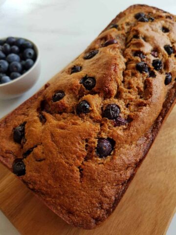 Banana bread with blueberries on a wooden board with a bowl of blueberries on the side.