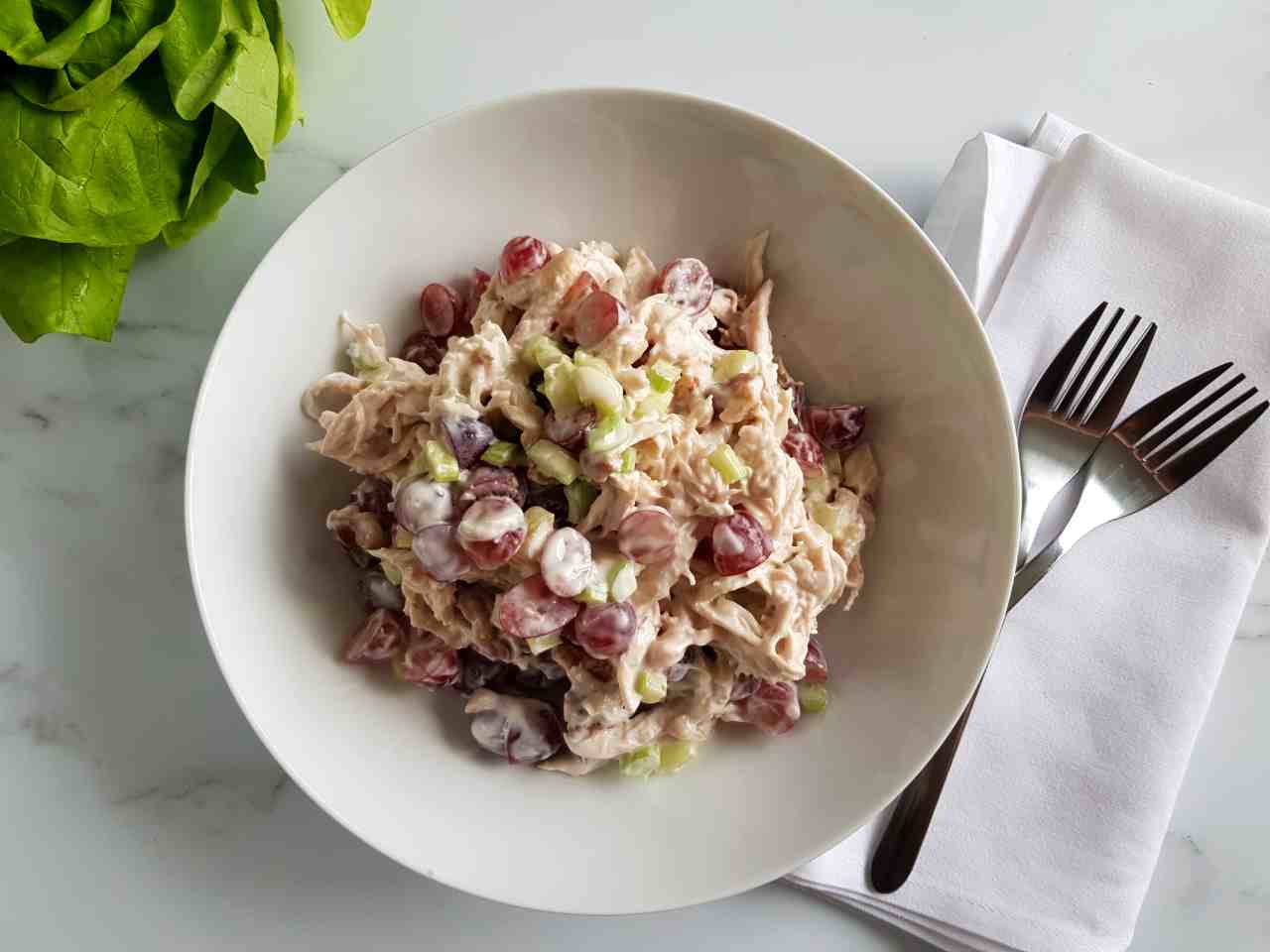 Grape and chicken salad in a white bowl on a marble table. Forks places on a white napkin on the side.