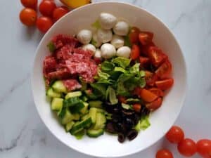 Italian salad in a white bowl on a marble table with cherry tomatoes scattered around.