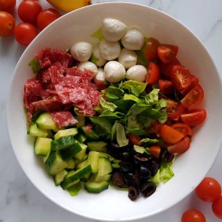 Italian salad in a white bowl on a marble table with cherry tomatoes scattered around.