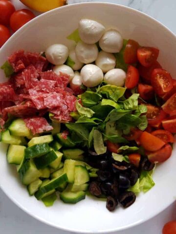 Italian salad in a white bowl on a marble table with cherry tomatoes scattered around.