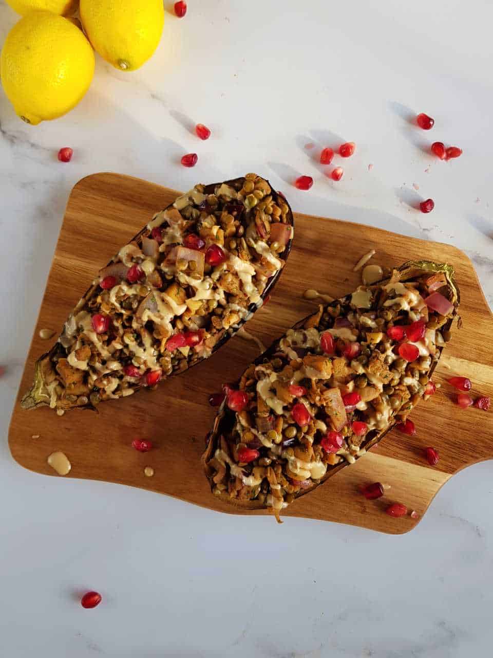 Stuffed aubergines with lentils on a wooden board with pomegranate seeds scattered around.