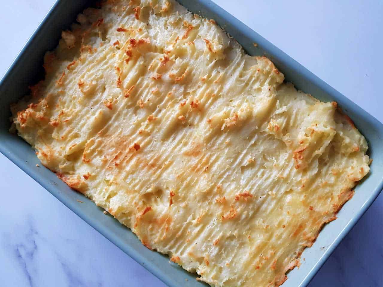 Vegetarian cottage pie with lentils in a green casserole dish on a marble table.