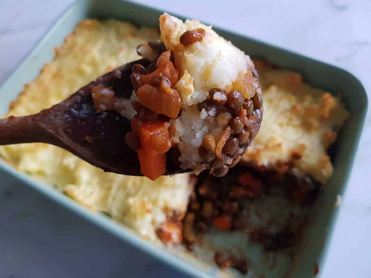 Vegetarian cottage pie in a green casserole dish. A wooden spoon has lifted some of the dish closer to the camera.