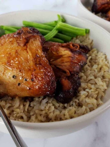 Adobo chicken in a white bowl with rice and green beans on a marble table.