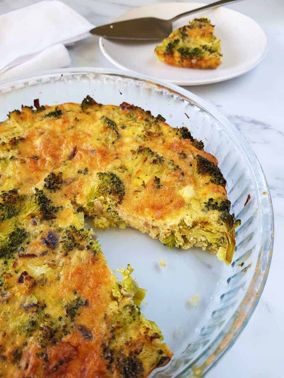 Broccoli quiche with no crust on a white table with a plated slice in the background.