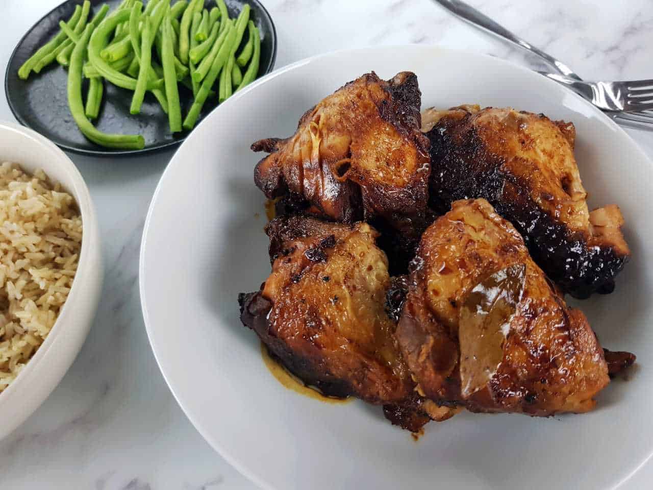 Chicken adobo in a white bowl with rice, green beans and cutlery on the side.