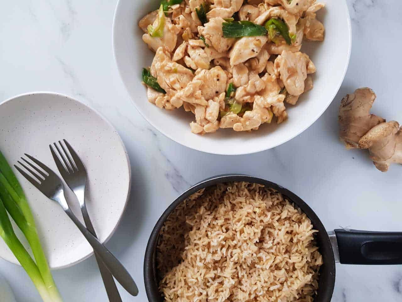 Chicken with ginger and spring onion in a white bowl with a pot of rice and spring onions on the side.