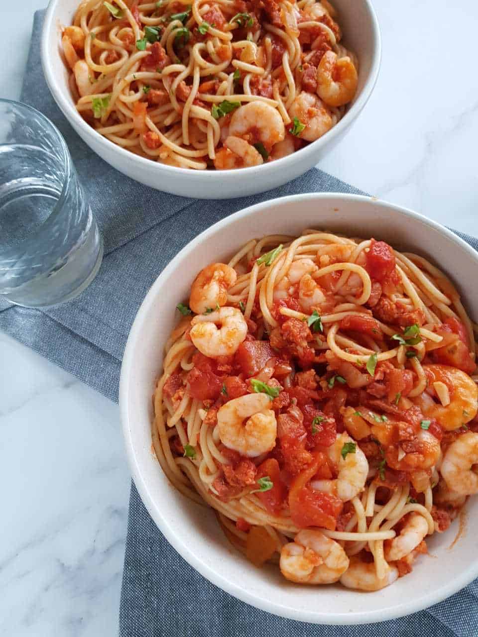 Chorizo and prawn pasta in white bowls on marble table with glasses of water on the side.