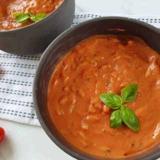 Creamy tomato soup with orzo in gray bowls on a white table with cherry tomatoes on the side.