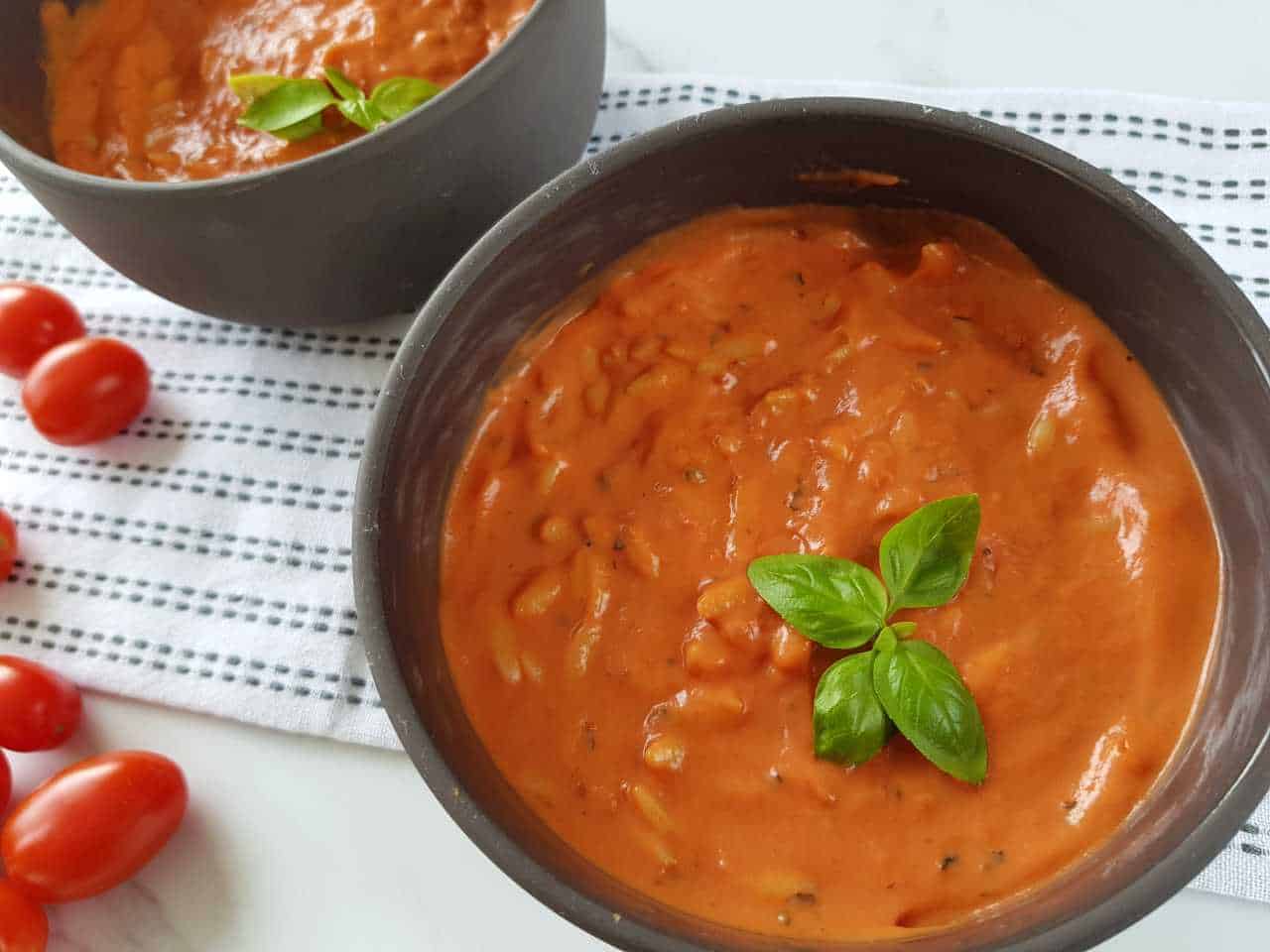 Creamy tomato soup with orzo in gray bowls on a white table with cherry tomatoes on the side.