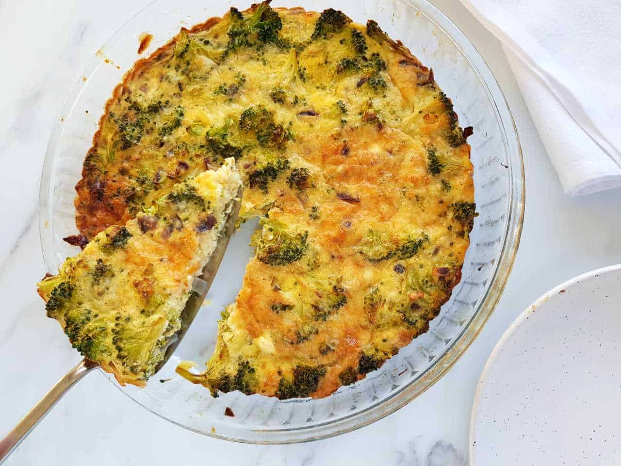 Crustless broccoli quiche on a white table, a cake server lifting up a slice.